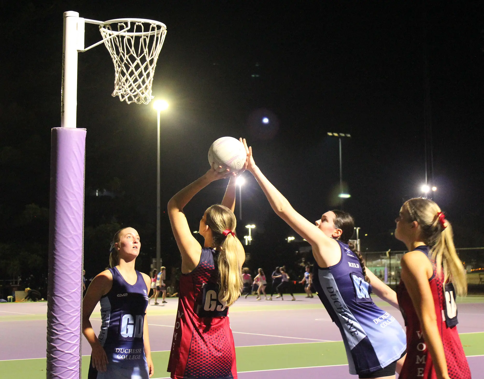 Four netball players from two teams, with one person aiming to score a netball goal.
