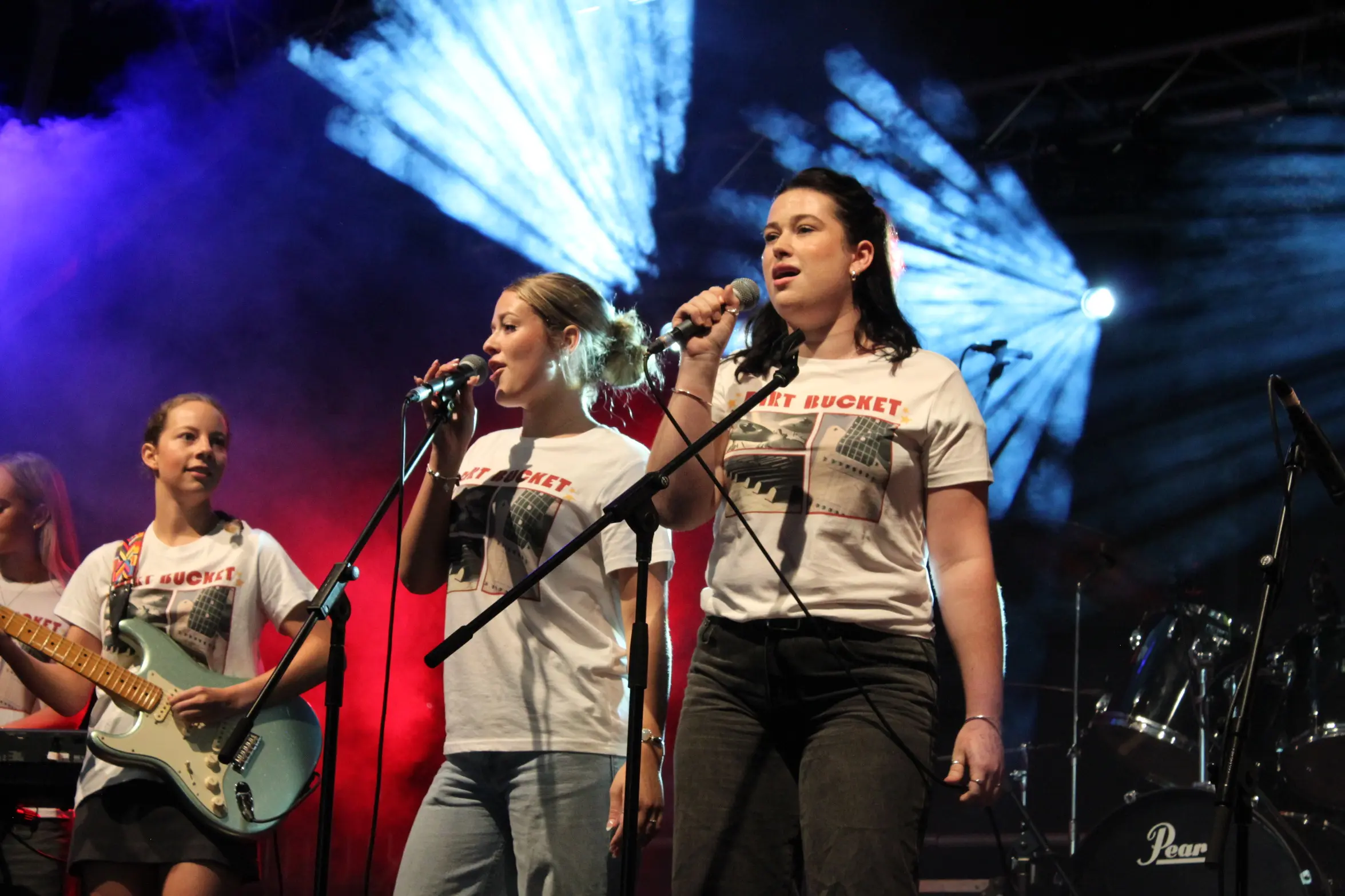 Three students perform in a band, with two singers and one guitarist visible.