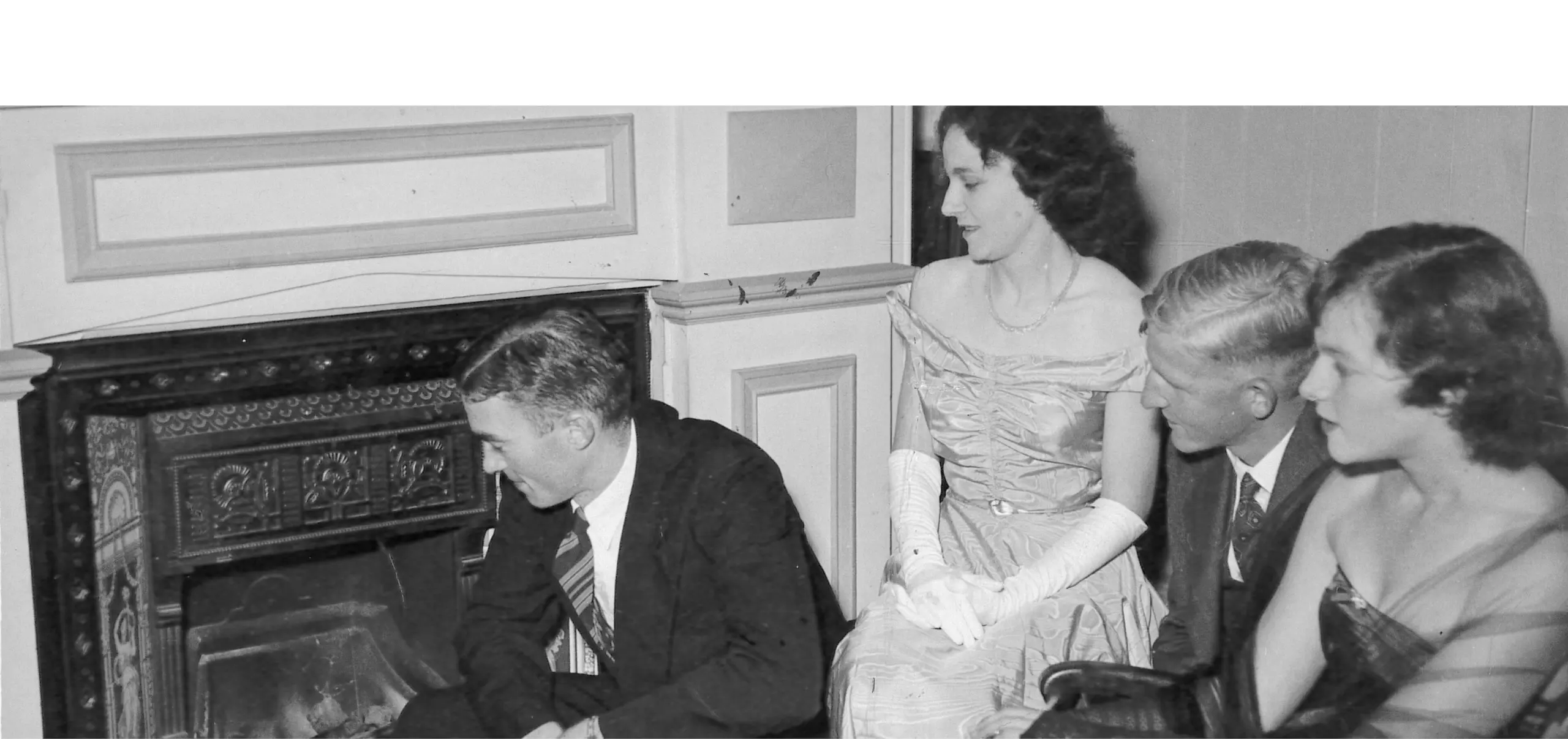 A black and white photo of a group of people in formal dress in front of a fireplace.