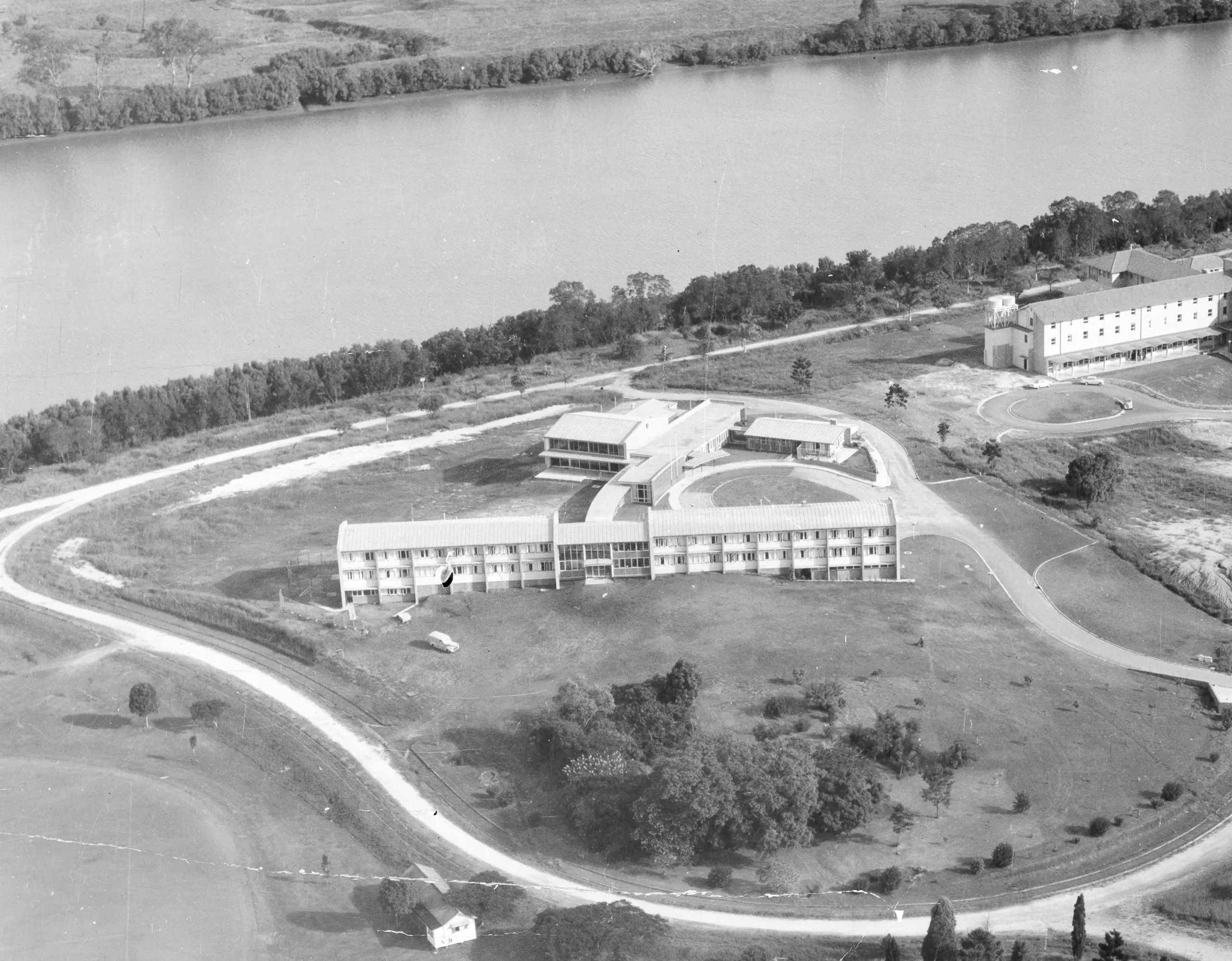 A black and white aerial photograph of three buildings surrounded by empty land and a river towards the back.