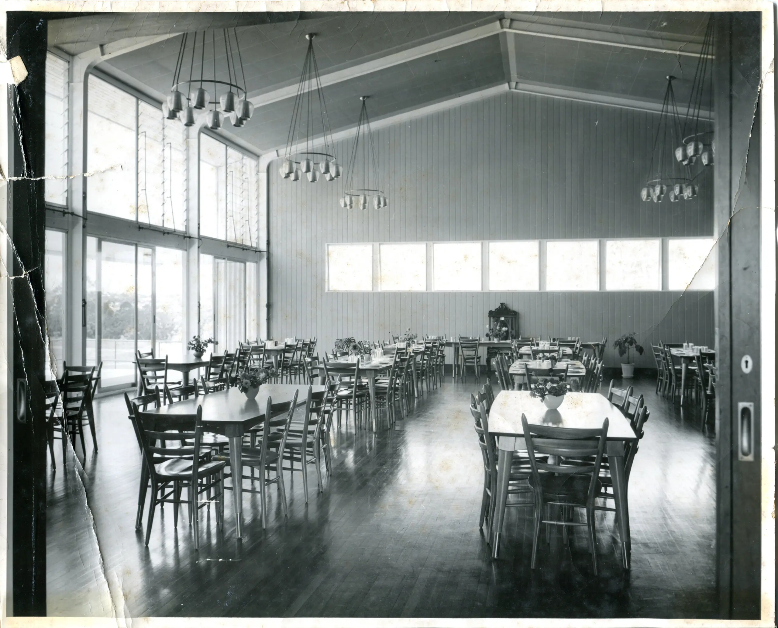 A black and white photo of the inside of a dining hall, with tables and chairs on the floor.