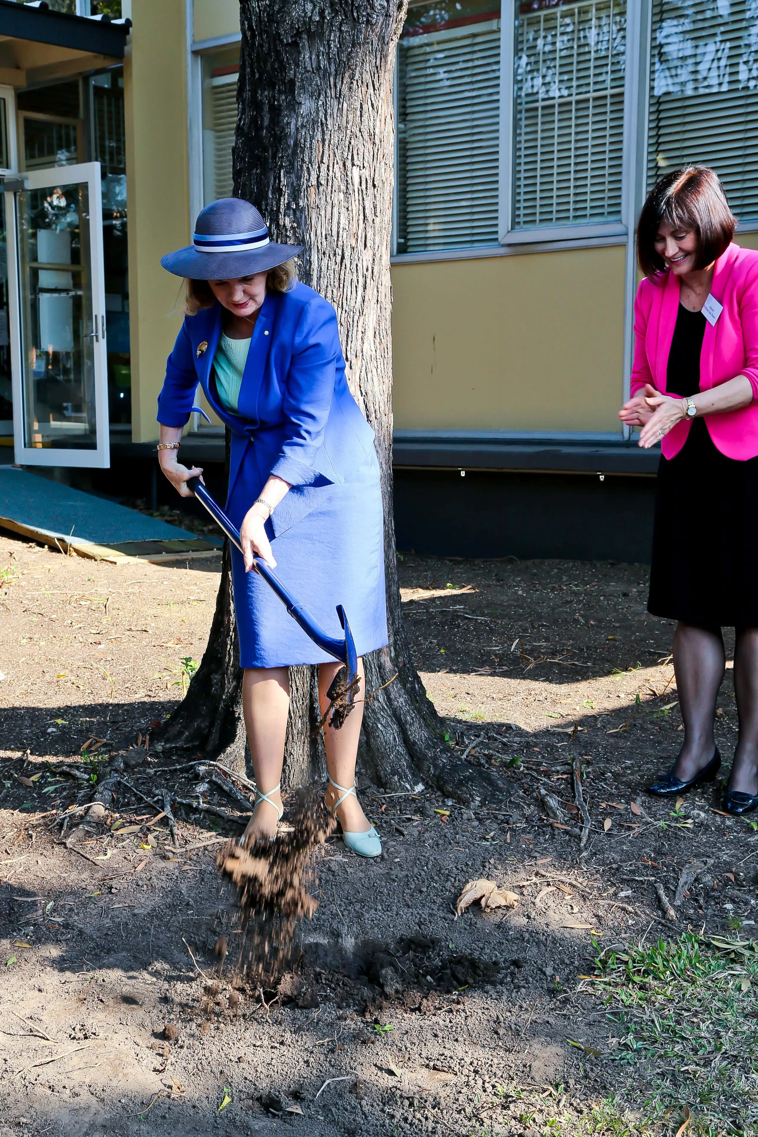 A woman in a blue suit and blue hat turns the sod in front of a tree, and is watched by a woman in a black dress and pink jacket.