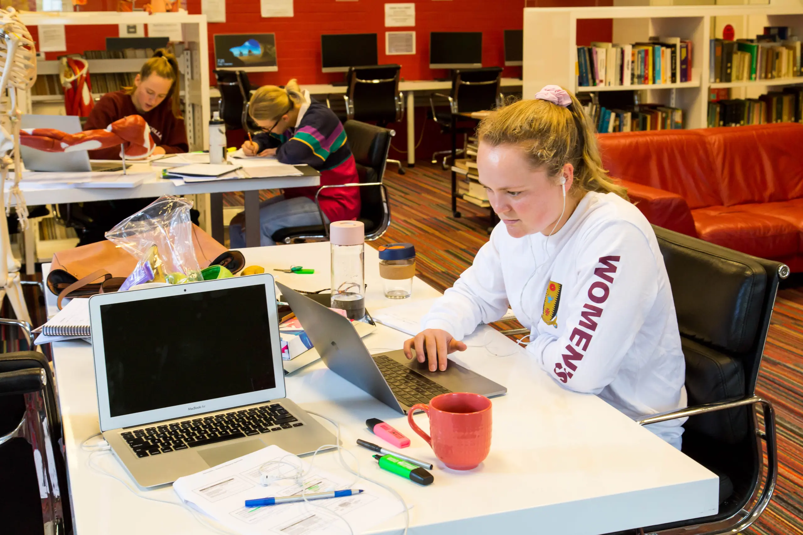 A student uses a computer in the foreground, while in the background you can see more students studying. The photo is an interior image of a library.