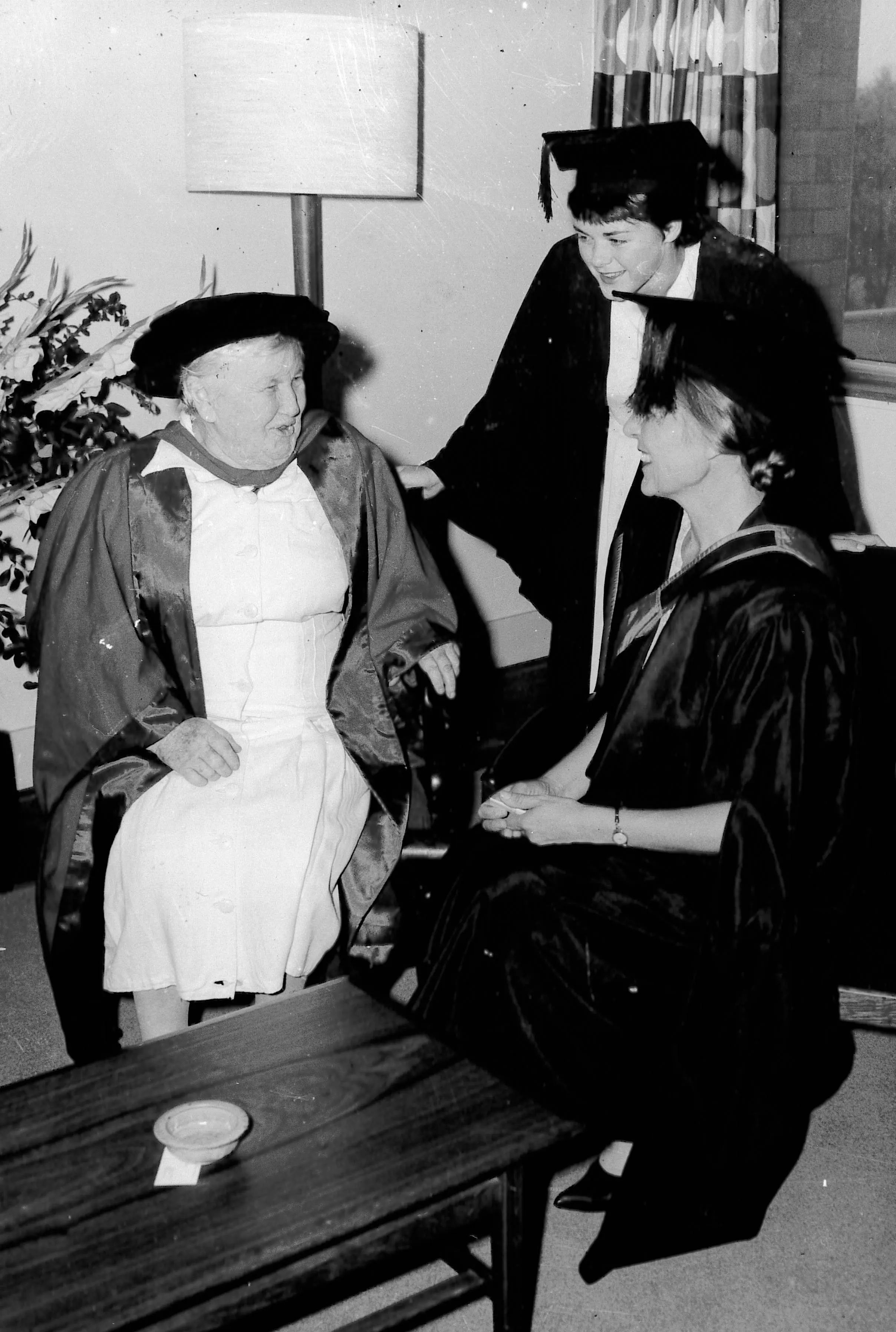 A black and white photo of three women wearing academic gowns and hats.