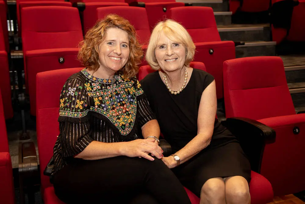 Two women sit in chairs in a lecture theatre.