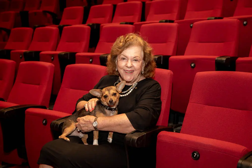 A woman sits on a chair in a theatre. She has a dog on her lap.